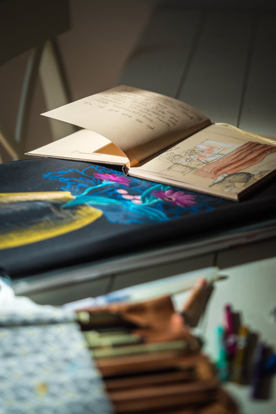 A drawing on black paper, a sketch book and some art supplies on a table, illuminated by morning light