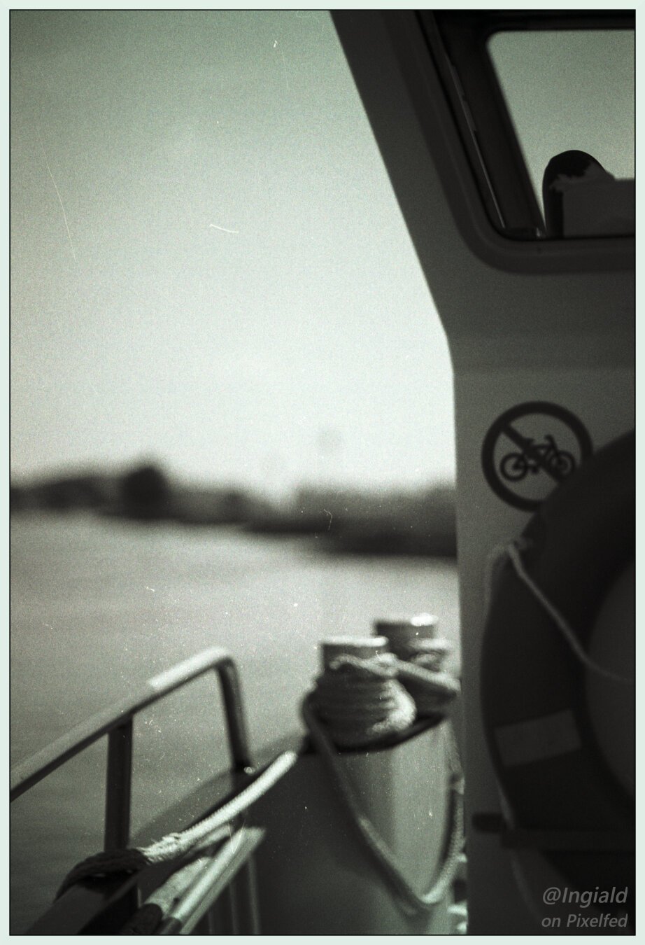 Black and white picture taken from the ferry. With the wheelhouse on focus on the right of the frame and the river out of focus on the left. The wheelhouse holds a lifebuoy and a sticker prohibiting bicycles. The railing of the ship is visible in the lower third of the frame with some bollards holding ropes.
Shot with a Minolta XD-7 with 85mm 1.7. on TMAX 100 developed in Rodinal 1+50.