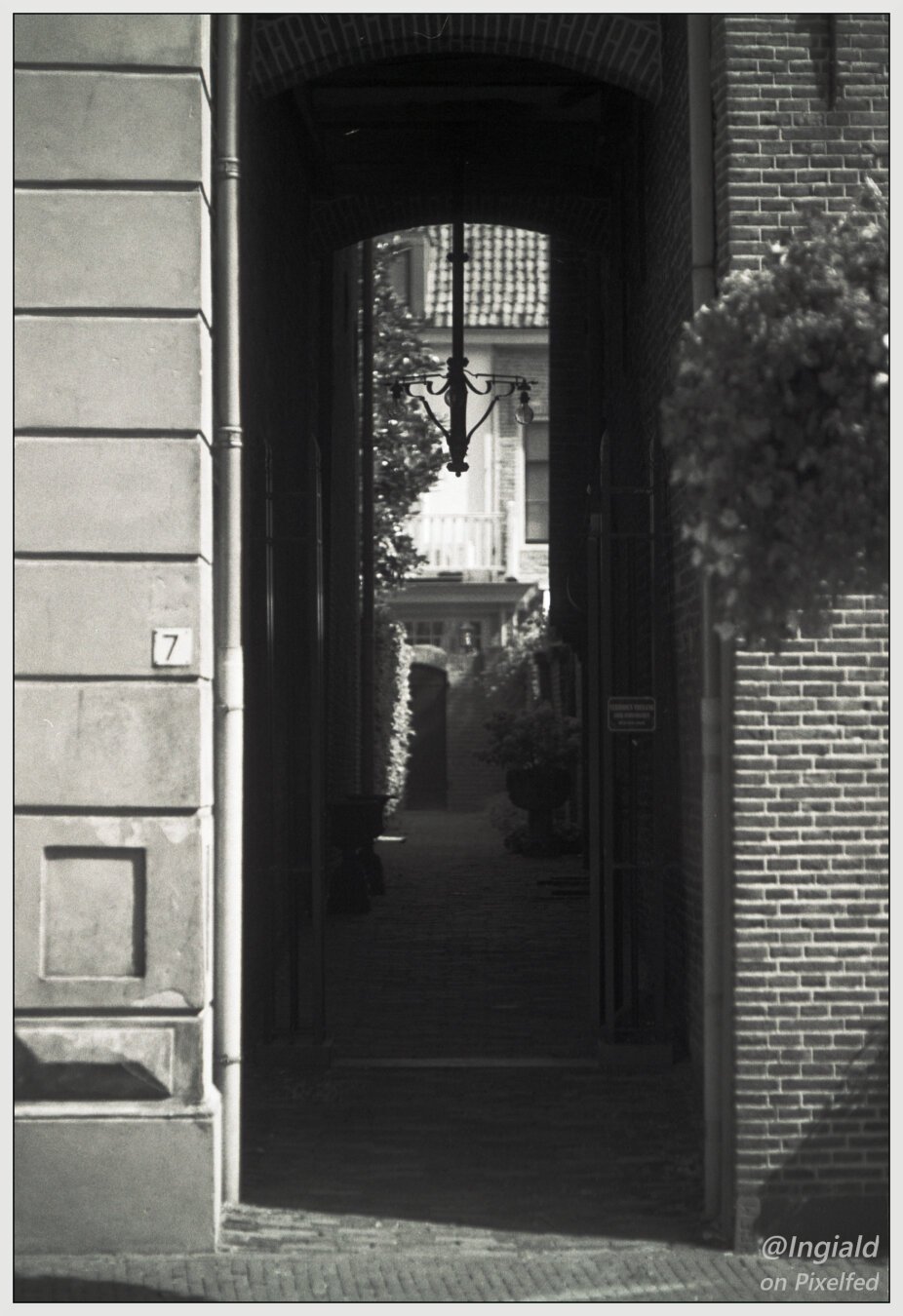 Black and white photograph of an alleyway leading to a door in a small courtyard. In the alleyway hangs a small iron chandelier.