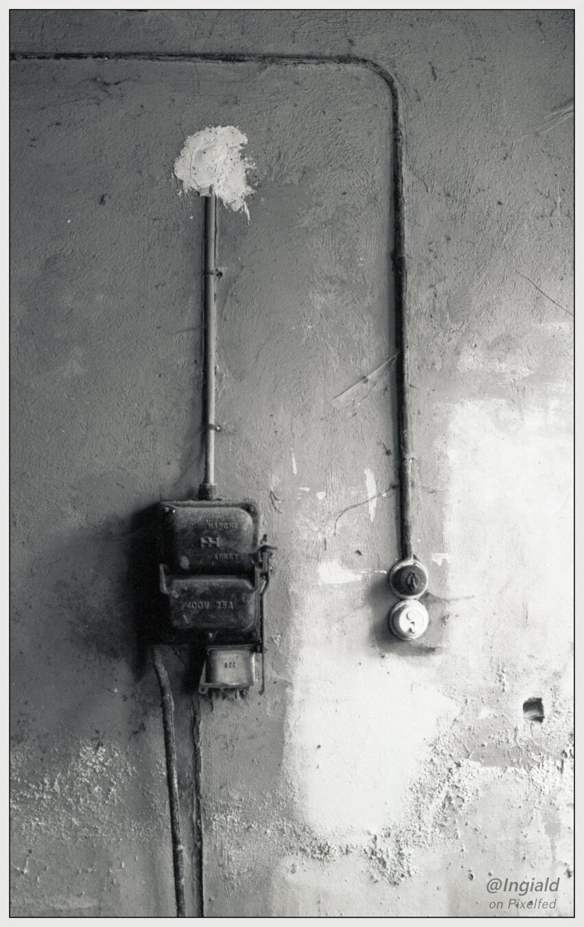 Black and white picture of a wall holding a big electrical switch. It says: MARCHE ARRET and 400V 25A. The power lines running along the wall half submerged in stucco. Next to the big switch are two ordinary light switches.