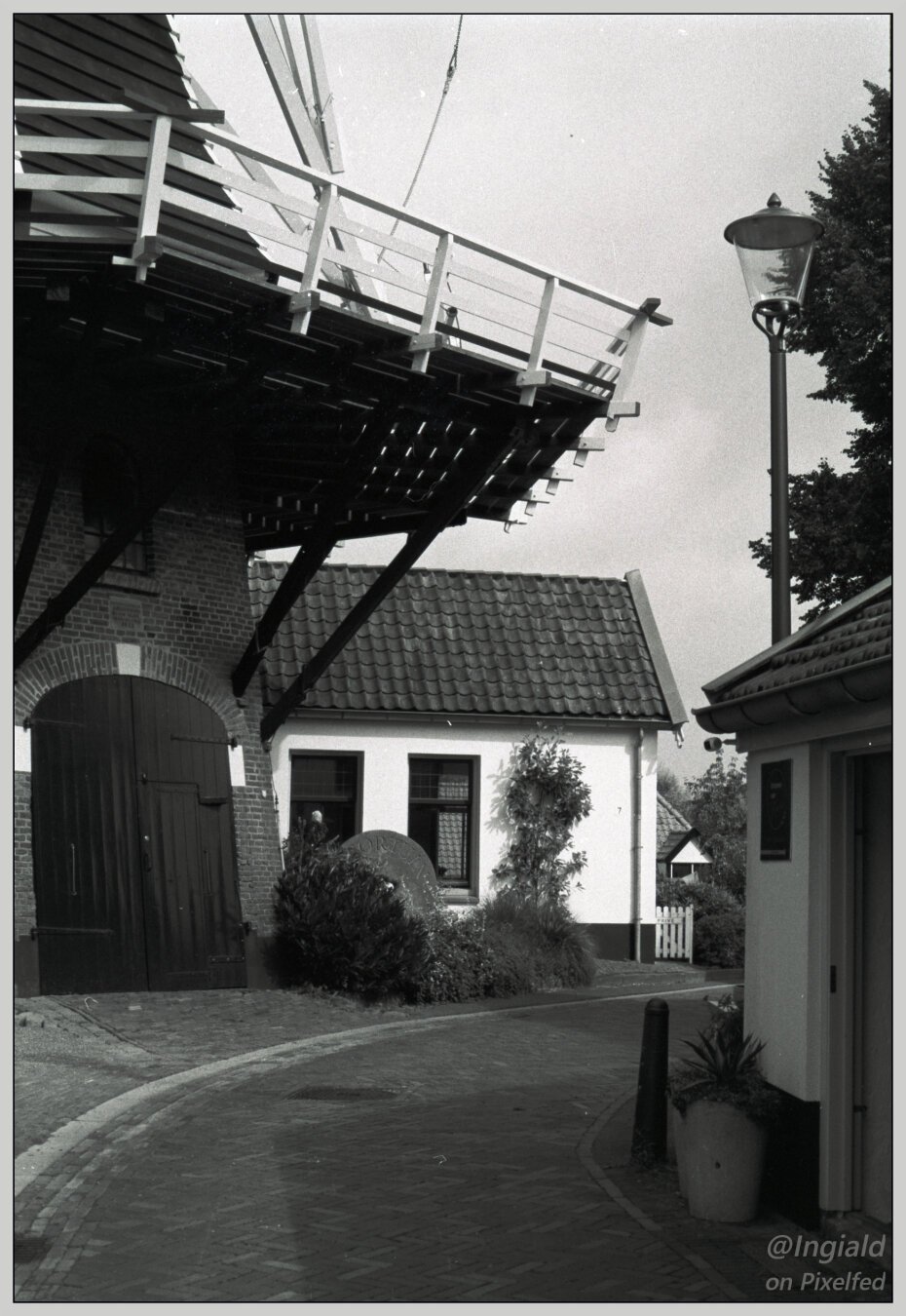 A black and white photograph of a bend in a cobbled street running right next to a windmil. The brick base of the mill holds big double doors, above it looms a big wooden plateau with balustrade spanning over the street below. More buildings abut the mill and the street either side. One of the old milling stones has been set upright in a patch of greenery nearby.