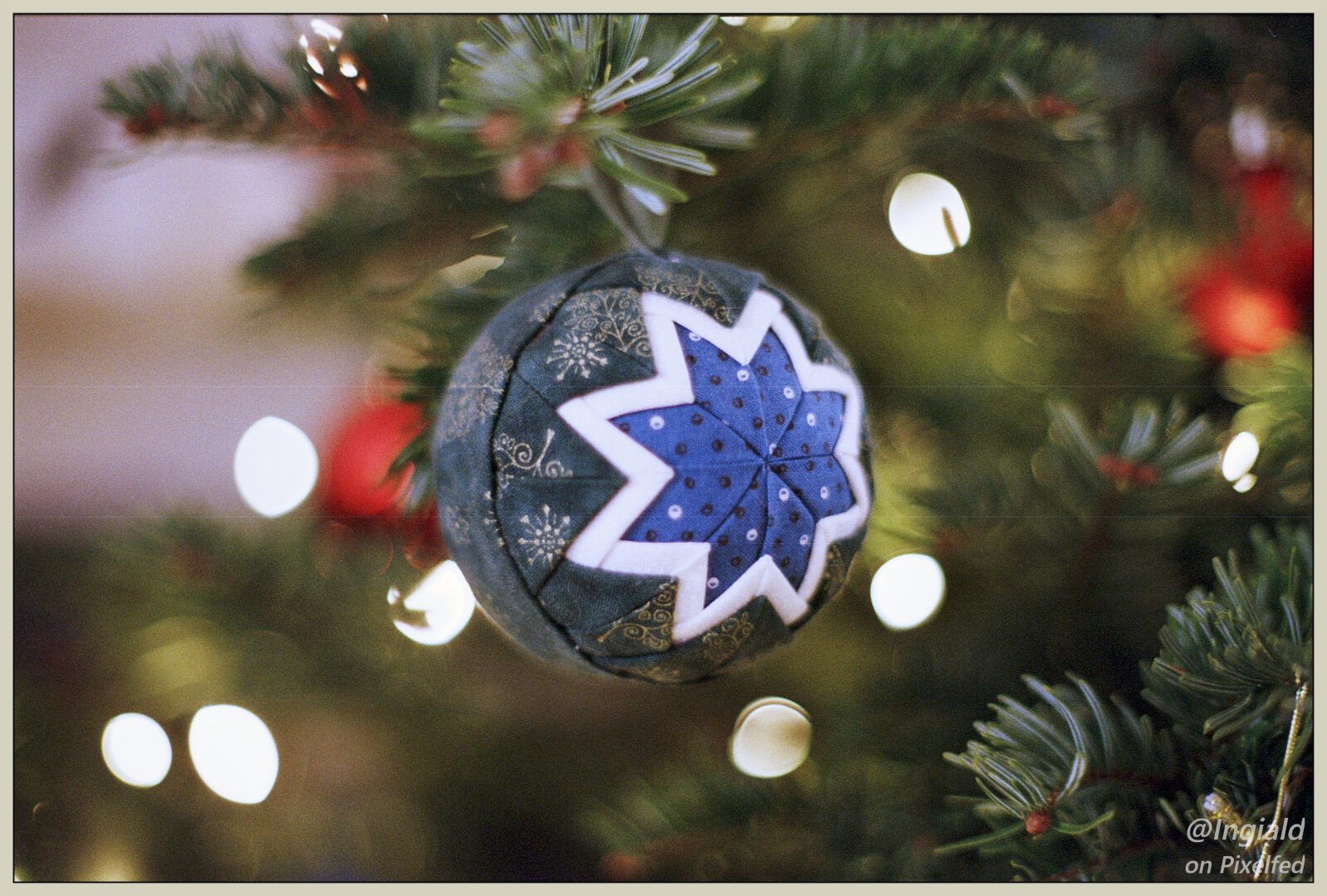 A colour photograph of a Christmas bauble hanging in a Christmas tree. The ornament is made of quilt fabrics in a star shape lain over a sphere. The background is the tree and some lights out of focus.