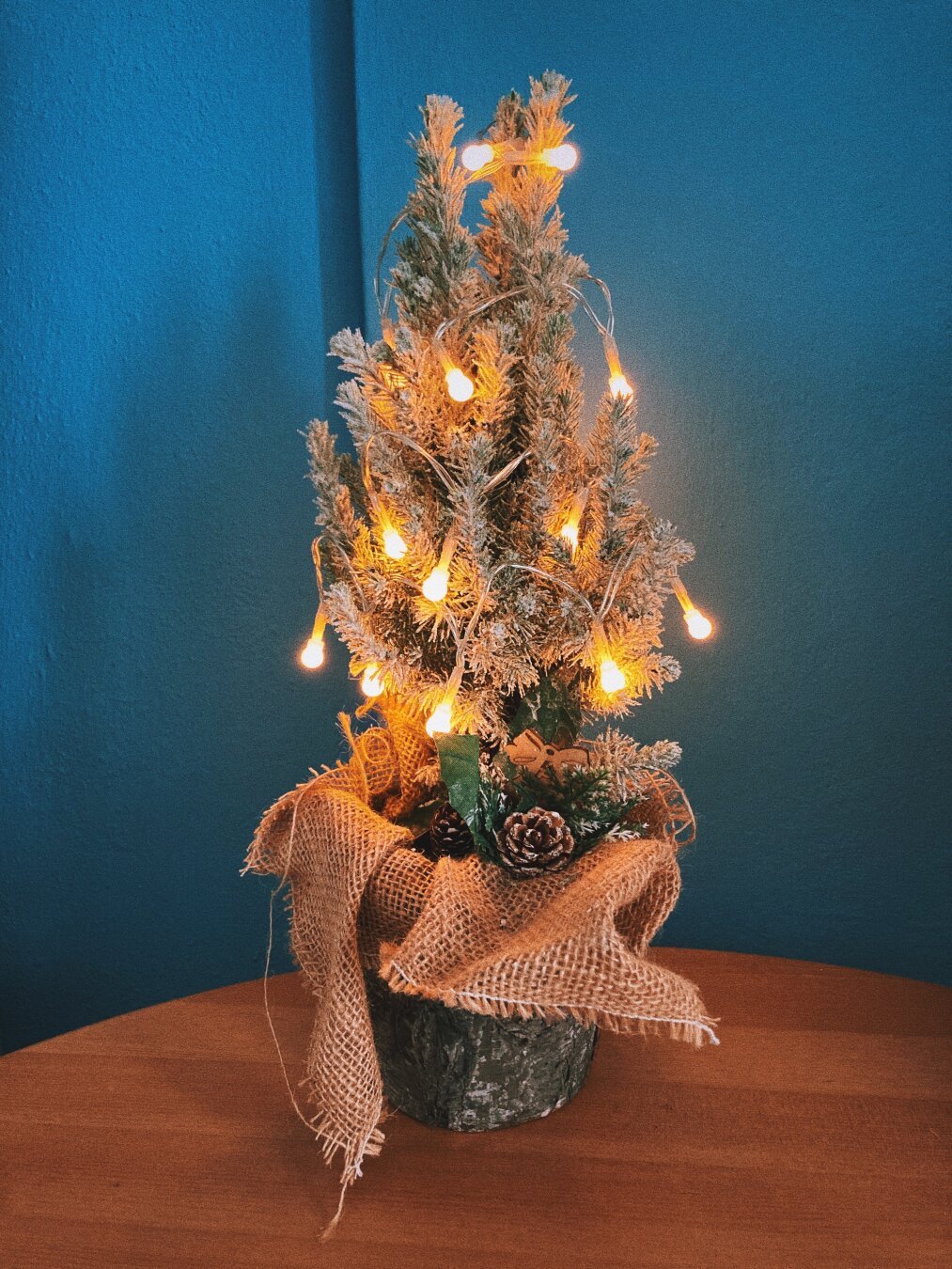 A small Christmas Tree with artificial snow and warm yellow LED lights, standing on a wooden table against a blue wall.