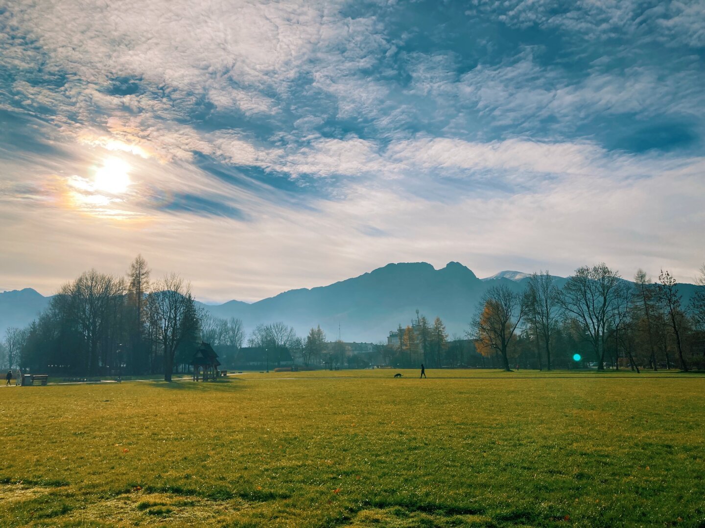 Late evening hours. Mountain range seen in the distance, partially covered by haze. In the foreground large grass area with some trees at the edges. Sun is visible fairly low on the horizon, partially covered by clouds.