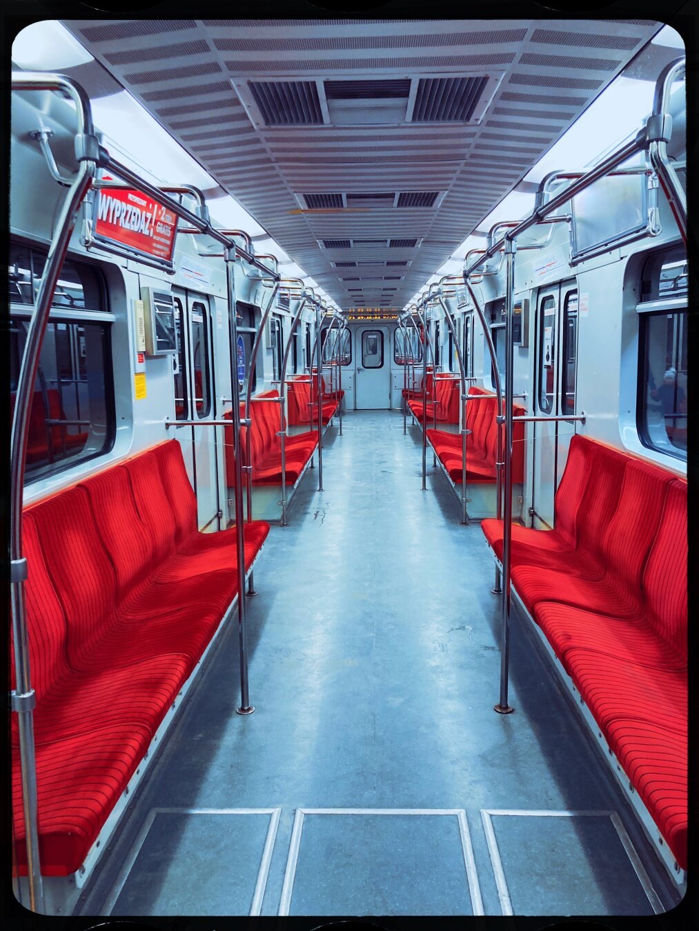 An empty carriage of an underground city train. On both sides there is a row of empty seats covered with intensive red fabric, floor has a glossy texture with a blueish / greyish hue. Carriage is well lit, exterior seen through windows is dark due to the train being in a tunnel.