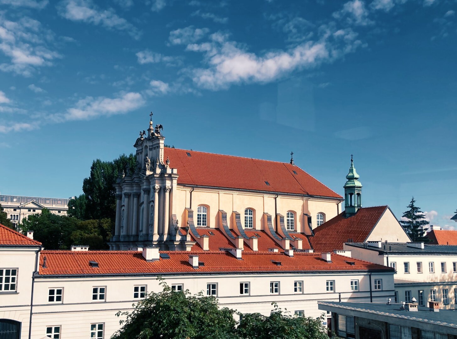 View from a window on the third floor, showing a cityscape with long white building in the foreground and a church in the background. All buildings have red roofs. It is a sunny day around noon. Sky has deep blue colour with few white clouds.