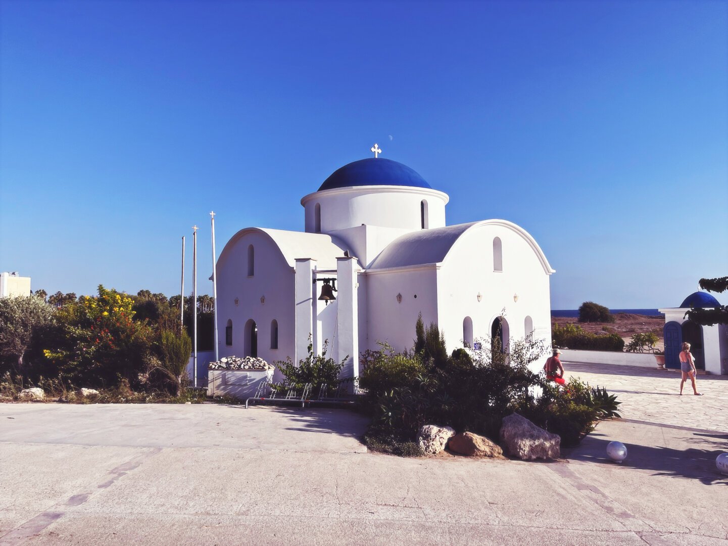 Small Greek Catholic church seen from a distance of a couple of meters, with white walls and blue roof in a shape of dome. There is low vegetation around, in form of palms and bushes. Deep blue sky is clear. Sun is not visible but is casting shadows from the far right. Late afternoon.