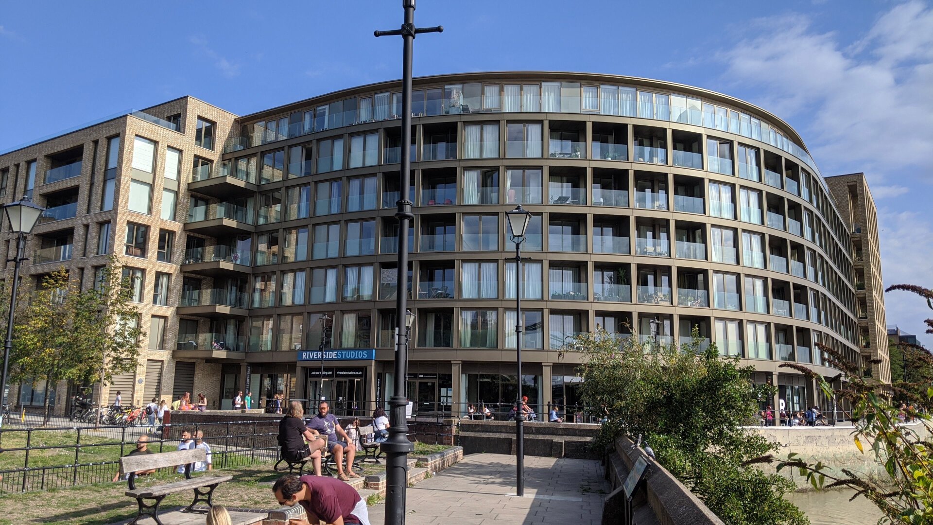 A 7 storey modern building with a rounded corner presenting windows and balconies for the flats which form the upper floors. Over the doorway on the ground floor a sign says Riverside Studios. in the foreground people enjoy the sunshine on a riverside walkway.