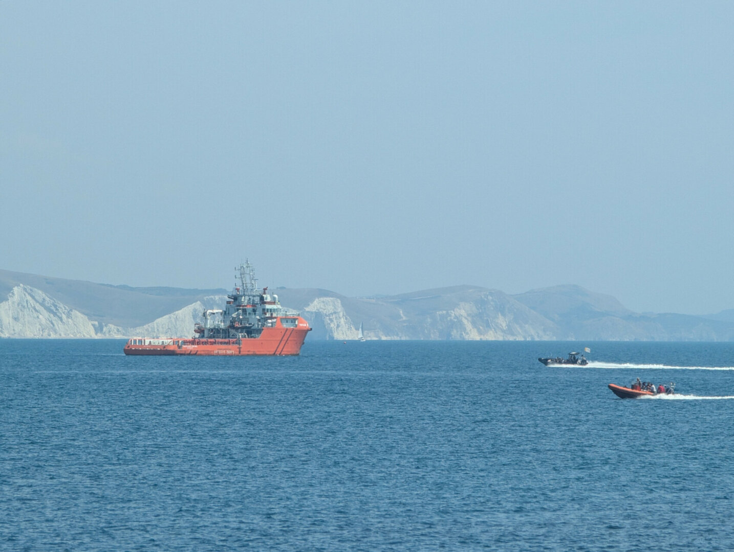 An orange ship anchored in bright sunshine on a calm sea. A line of white cliffs is in the background. In the foreground two small fat boats speed across the water from the right, trailing white wakes