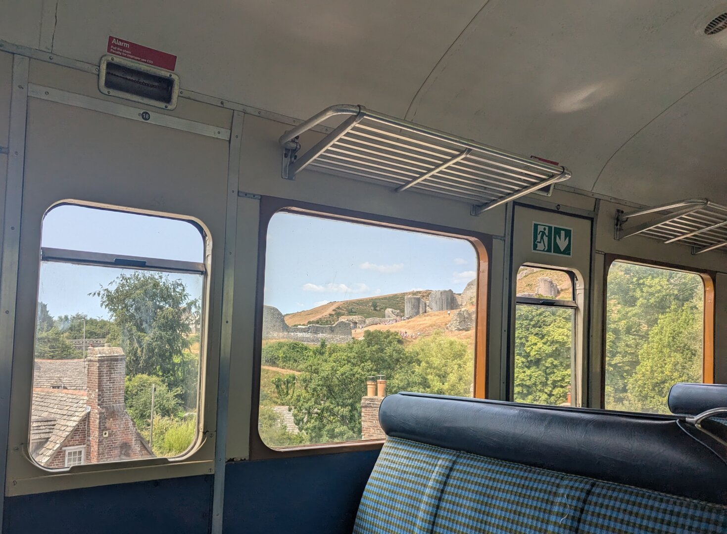 Looking out through the windows of an old train carriage. The train is raised on an embankment so through the windows we can see roofs and chimneys, trees, and on a hill the ruins of a castle