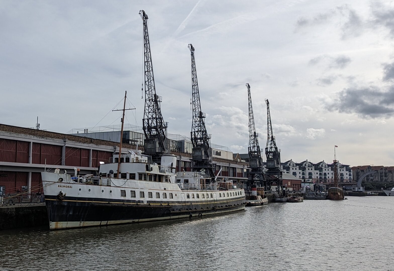 A small black and white passenger ship moored alongside a city wharf with a large warehouse and four very tall cranes behind it