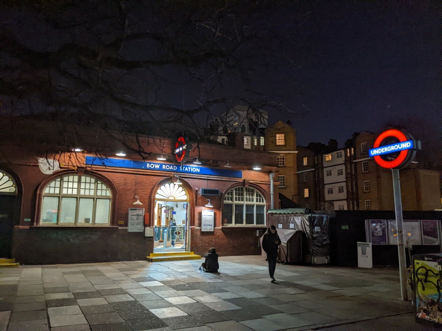 Night time photo of a red brick fronted building with an arched doorway flanked by two arched windows. A white on blue sign above the door gives the station name, with a projecting Underground roundel above it. Another internally lit roundel on a totem pole is on the right in the foreground. Light floods from the doorway onto the paving stones of the forecourt illuminating a beggar sitting facing the doorway and a second person who has walked out of the station passing the beggar.