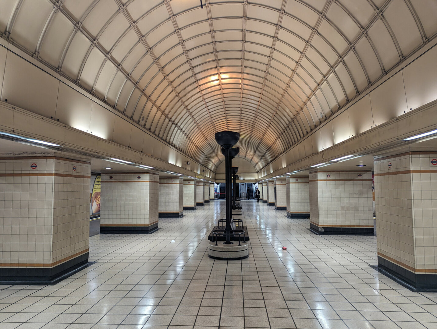 In an underground station looking along a passenger circulating space with stout square tiled columns on each side supporting a high arched ceiling. along the middle are tall hemispherical up lighters with back to back seats on the plinths at their bases.