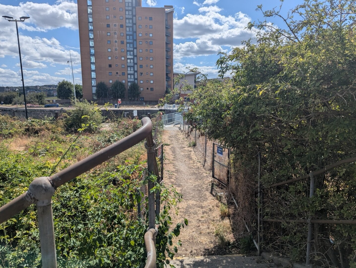 A path leads straight ahead between wire mesh fences. It passes through a pair of open gates. On the left is scrubby wasteland, to the right bushes overhang the fence. At the end of the path a large set of lock gates is visible over which the path continues. Beyond the lock is a tall block of flats.