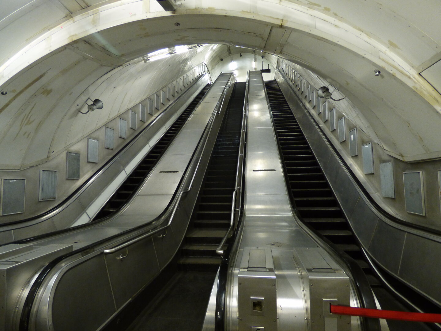 two shiny stainless steel escalators with a staircase between them lead up through an arch roofed tunnel. Poster frames for advertisements line the walls on both sides but are all empty. The scene is brightly lit by strip lights above