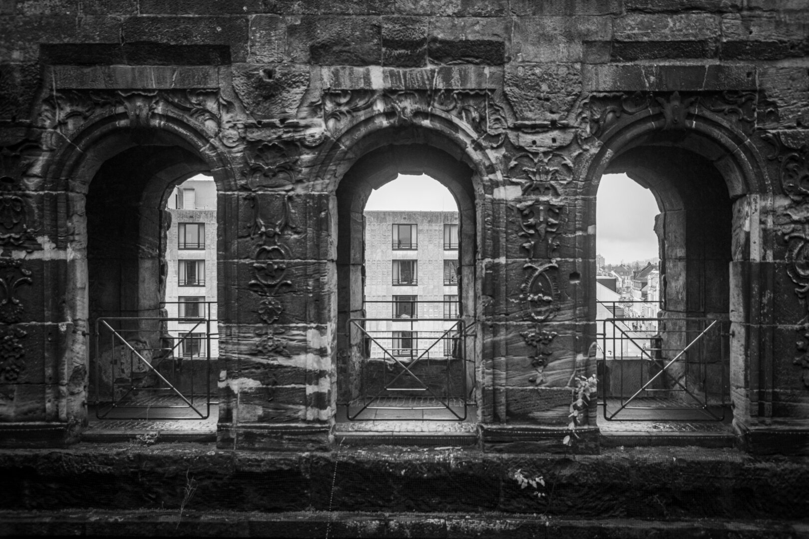 a photograph of the inner part of Porta Nigra. You can see the black fascade and three windows. Through the windows you can see the fascade of a modern building