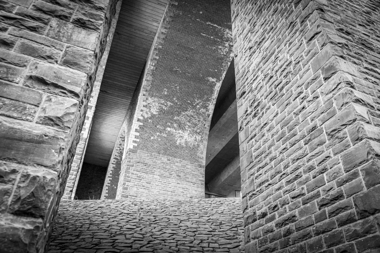Schwarz-Weiß-Foto einer Talbrücke von unten. Die Brücke besteht aus hohen Steinmauern mit unregelmäßigen, groben Steinen. Im Hintergrund sind ein Metallgitter und die Unterseite der Brücke zu sehen. Der Boden ist mit unregelmäßigen Steinplatten gepflastert. Die Perspektive ist nach oben gerichtet.