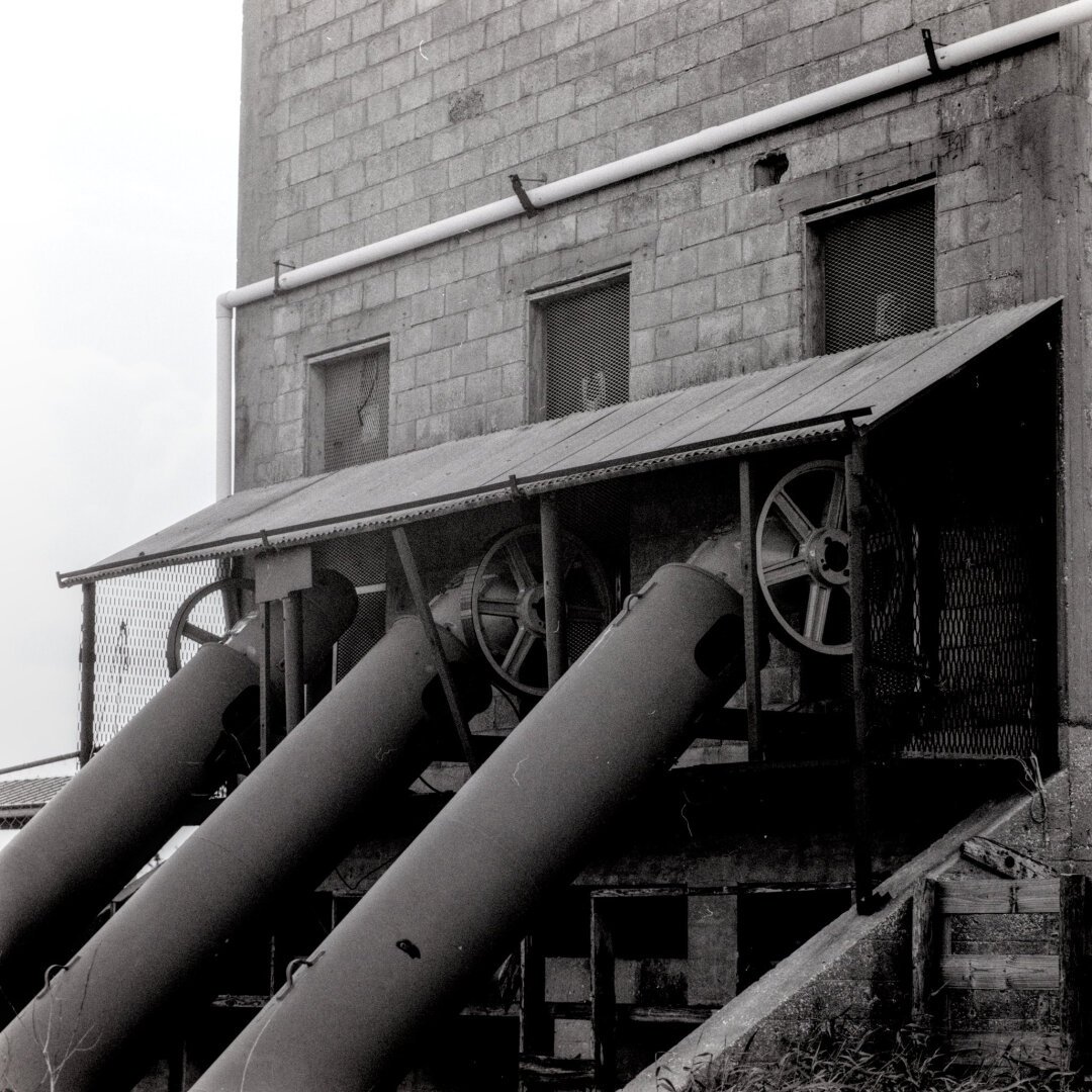 A black and white photo of a large old pumphouse. Three large pipes extend off to the side of an old brick building.