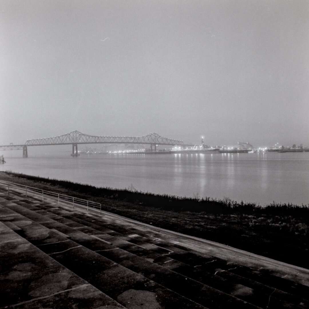 A black and white photo of the Mississippi river taken from the east bank and looking towards barges and a bridge.