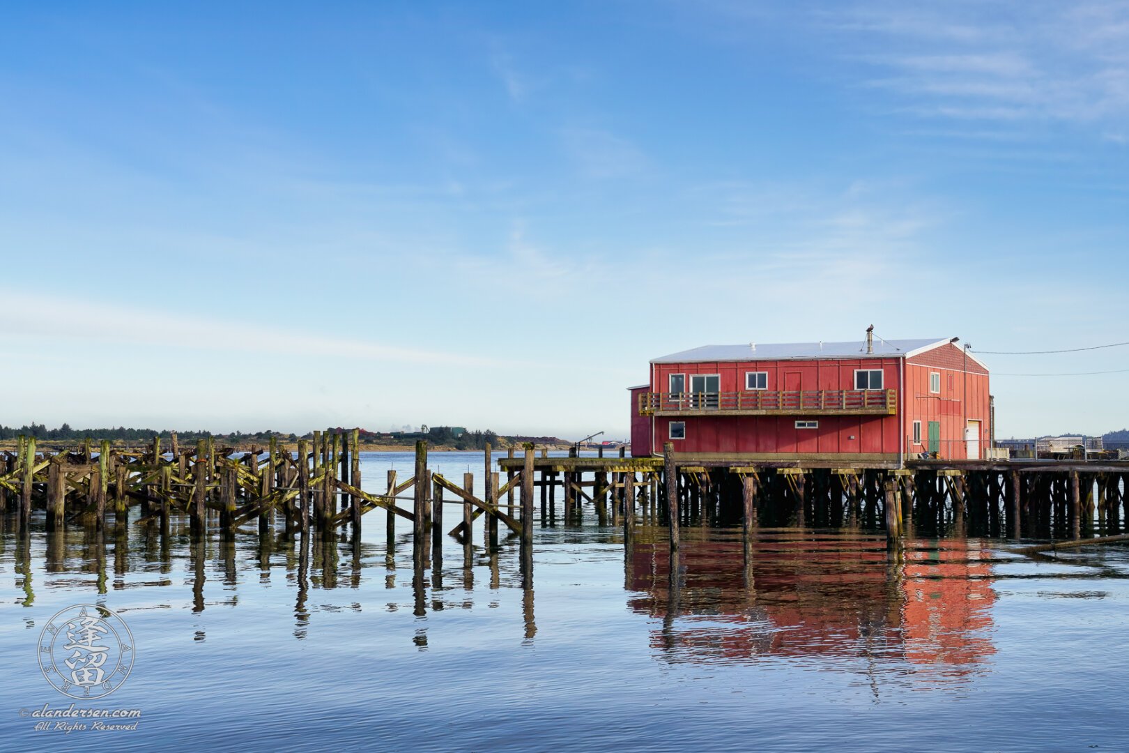 A red two-story building sitting on a pier, lit by the morning light against a bright blue sky with soft streaky clouds, and surrounded by the posts of old piers that have long since fallen into disuse and the waters of the river that reflect this imagery.