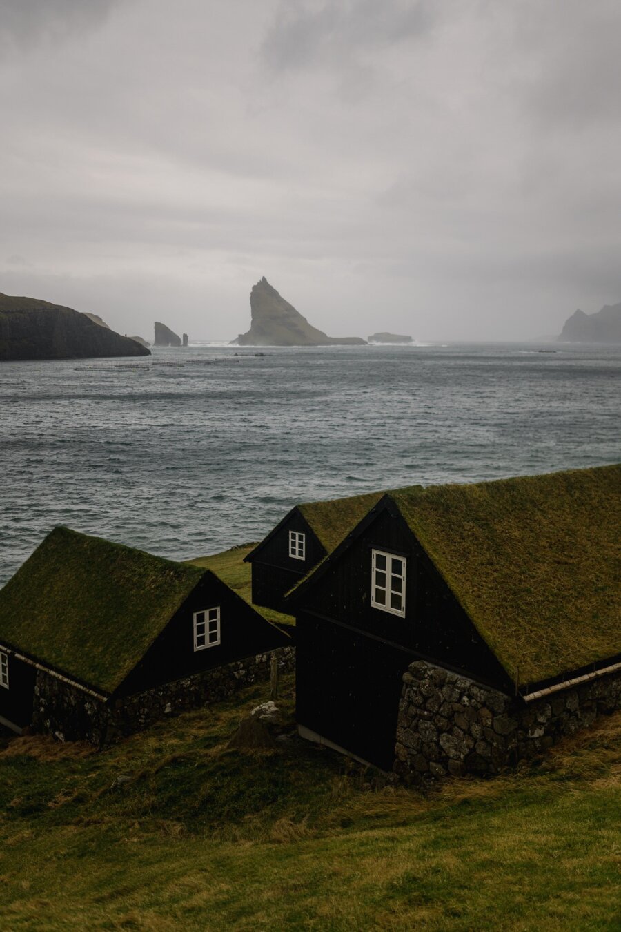 This image captures a moody coastal scene in the Faroe Islands, featuring Tindhólmur, a striking island with sharp peaks rising from the sea. In the foreground, three traditional Faroese houses with black wooden walls and grass-covered roofs sit on a sloping hillside. Their stone foundations blend into the rugged landscape.

Beyond the houses, the choppy, deep blue-gray sea stretches to the horizon. The misty cliffs and smaller rocky islets surrounding Tindhólmur add to the remote, untamed feel. Heavy, low clouds cast a soft, diffused light, enhancing the cool, muted tones of greens, grays, and blacks. This scene perfectly embodies the wild beauty of the Faroes.