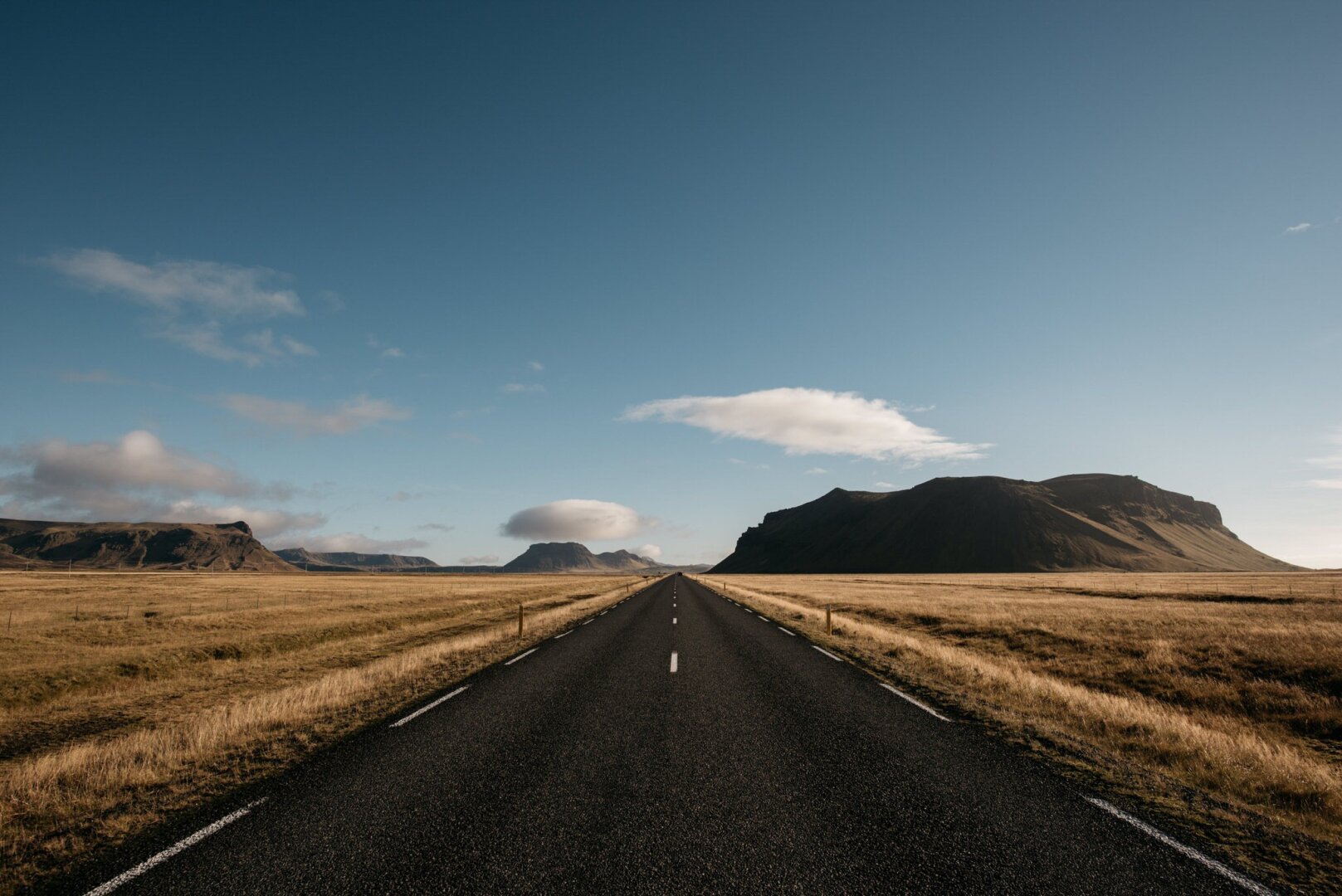 The image depicts a long, straight road cutting through a vast, open Icelandic landscape. The road is bordered by flat, golden-brown fields of grass stretching into the distance. On either side, there are dark, rugged mountains with gently sloping shapes, highlighted by soft light. The sky above is expansive, with patches of soft blue and scattered white clouds, creating a sense of calm and openness. The perspective of the road draws the viewer's eye toward the horizon, giving a sense of infinite possibilities and peaceful isolation in nature. It was taken bei European elopement and wedding photography Sturmsucht in October during a road trip with a wedding couple.