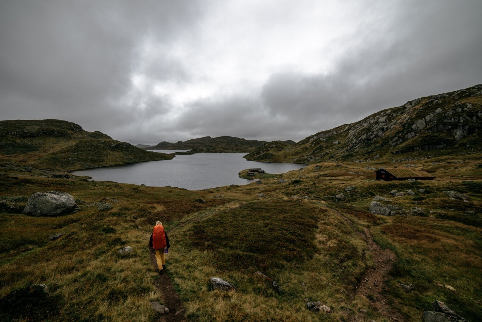 The image, taken by European elopement and wedding photographer Sturmsucht from Hamburg, depicts a remote, rugged landscape under a cloudy, overcast sky. The terrain consists of rolling hills covered in green and brown vegetation, suggesting a late summer or early autumn setting. A narrow dirt path winds through the scene, leading toward a calm lake surrounded by rocky slopes. In the foreground, a lone figure walks along the path, wearing a bright orange jacket and carrying a backpack, adding a pop of color to the otherwise muted palette. To the right of the lake, there is a small, dark wooden cabin nestled in the landscape, creating a sense of solitude and tranquility. The overall atmosphere feels moody and serene, evoking the feeling of exploration in a wild, untamed environment.