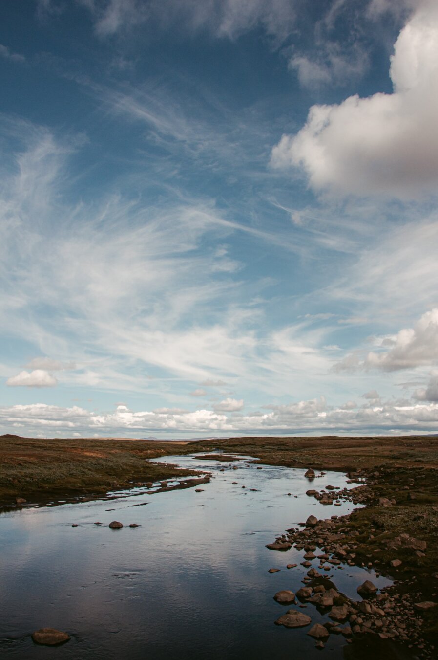 This image, captured in Iceland by European elopement Sturmsucht, portrays a vast and rugged landscape. A calm river meanders through a rocky valley, creating a sense of tranquility and stillness. Above, the sky is filled with soft, sweeping clouds that stretch across the horizon, adding a feeling of openness and movement. The riverbanks are sparse and raw, emphasizing the isolation and untamed beauty of the Nordic wilderness. The scene reflects a timeless and serene atmosphere, showcasing the connection between water, sky, and land.