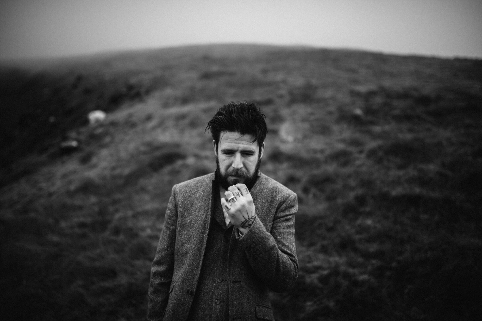 This black-and-white photograph captures a groom standing alone in a misty, open landscape. He is dressed in a textured, three-piece tweed suit, evoking a timeless and rugged style that blends seamlessly with the natural surroundings. His dark, slightly unkempt hair and full beard give him a thoughtful, introspective appearance.

His posture and expression convey deep contemplation—his brow is furrowed, and his gaze is directed downward. His right hand is raised to his mouth, fingers gently touching his lips, as if lost in thought. Tattoos are visible on his fingers and hand, adding a personal and artistic element to his presence.

The background is a vast, rolling hillside, covered in rough grass, fading into the mist. The atmosphere is moody and dramatic, with soft, diffused light creating a dreamlike quality. The lack of sharp background details draws attention to the groom as the focal point of the image by elopement und wedding photography Sturmsucht.