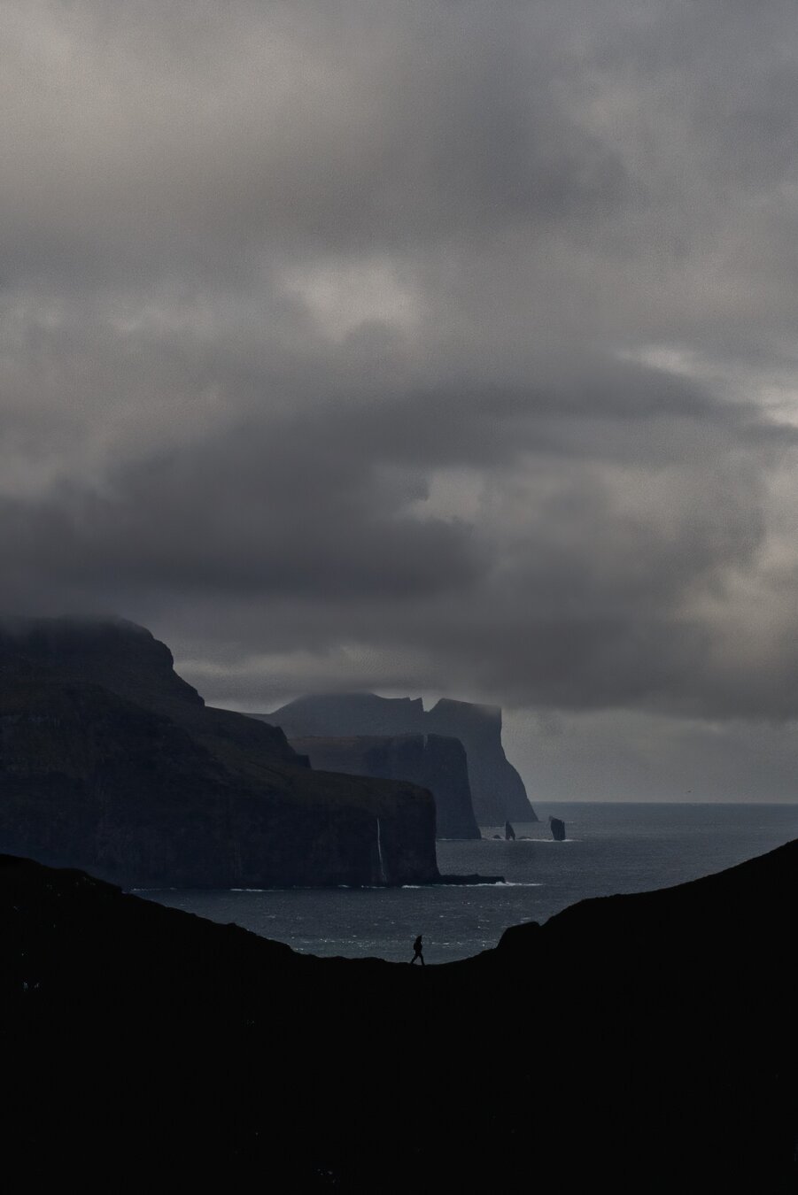 This is a striking and atmospheric landscape photograph taken by European elopement and wedding photographer Sturmsucht during an elopement on the Faroe Islands. The image captures a dramatic coastal scene under a heavy, moody sky filled with thick, textured clouds. In the foreground, a lone figure walks along a dark ridge, silhouetted against the stormy backdrop. Towering cliffs extend into the distance, with waterfalls cascading down their edges into the vast ocean below. The composition evokes a sense of solitude, adventure, and the raw beauty of untamed nature, perfectly aligning with Sturmsucht’s signature aesthetic of embracing wild landscapes and intimate human moments.