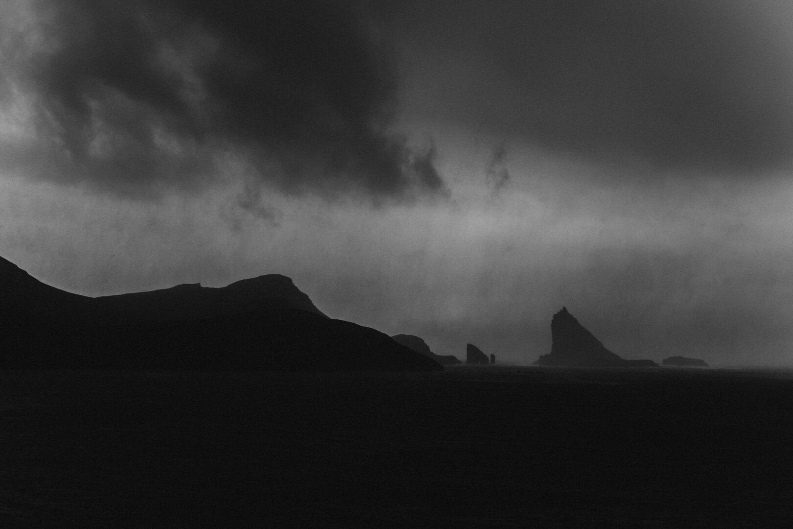 This is a black-and-white photograph depicting a dramatic seascape under a moody, overcast sky. Dark clouds dominate the upper portion of the image, creating a sense of intensity and movement. The silhouette of jagged rock formations rises from the ocean in the middle and right sections of the frame, contrasting with the softer lines of distant hills to the left. The absence of light and the stark contrasts create an atmospheric and mysterious feel, evoking solitude and raw natural beauty. The photograph captures a powerful interplay of shadow and texture, emphasizing the rugged landscape and the stormy weather.