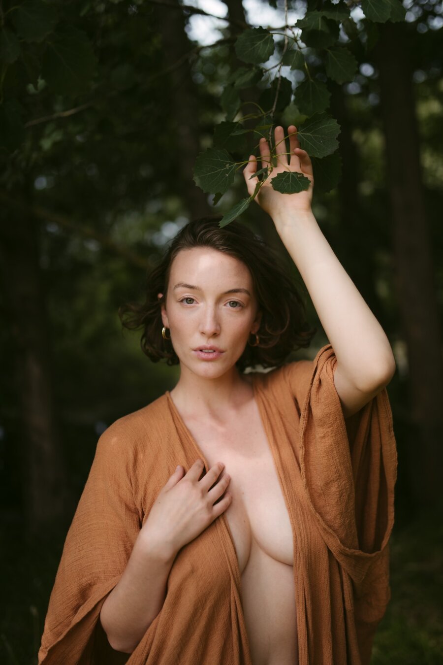 A woman stands in a sun-dappled forest, her gaze calm and direct. She wears a flowing, earth-toned robe that drapes loosely over her shoulders, revealing part of her bare skin. One hand rests gently on her chest, while the other reaches up to touch a branch with delicate green leaves. Her short, wavy hair frames her face, and golden hoop earrings catch the light. The background of blurred trees and soft shadows creates a natural, intimate atmosphere, blending her presence seamlessly with the surrounding wilderness.