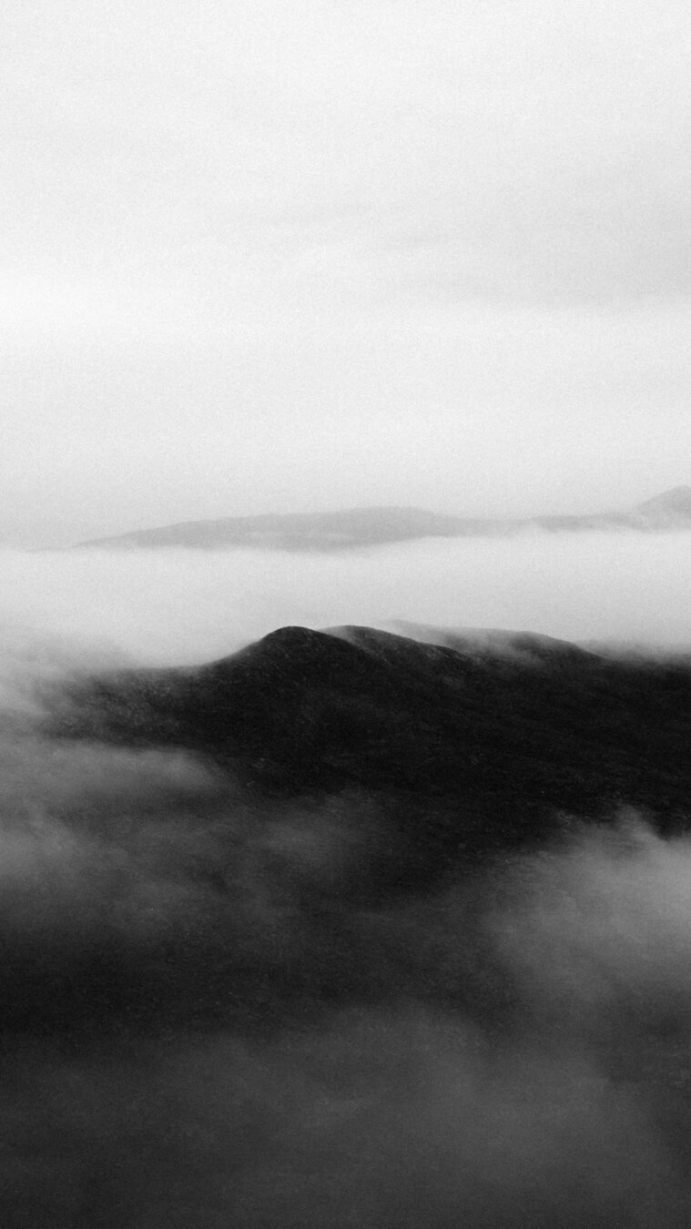 This image is a black-and-white photograph that captures a dramatic, misty landscape in Greenland. The scene is dominated by dark, rolling hills or low mountains partially shrouded in thick, swirling fog. The mist creates a soft, ethereal blanket over the terrain, obscuring some details while allowing the silhouette of the rugged land to emerge through the haze. The sky above is overcast and blends seamlessly with the fog, giving the entire scene a timeless, almost otherworldly feel. The contrast between the dark ground and the light mist enhances the stark, raw beauty typical of Greenland’s remote wilderness. The absence of color focuses attention on textures and contrasts, evoking a sense of solitude and quiet strength.