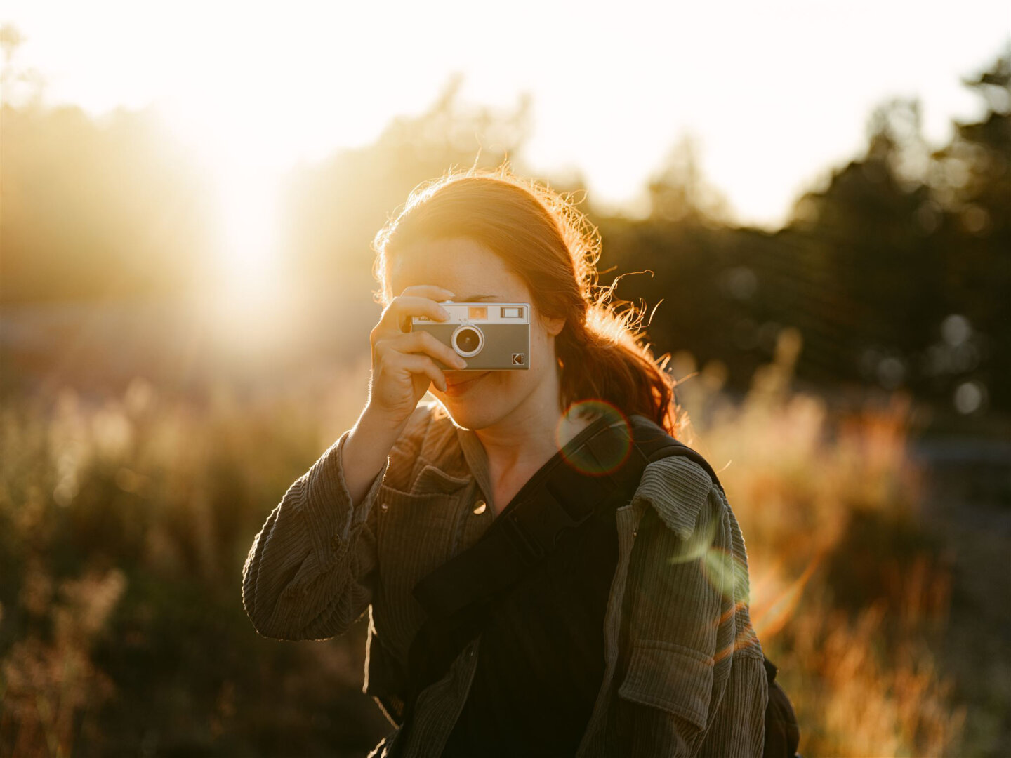 This image captures a person photographing with a vintage film camera during golden hour. The photographer has shoulder-length hair beautifully backlit by warm sunlight streaming from the left. They're wearing a dark olive corduroy jacket and holding a compact rangefinder-style camera with a silver body up to their eye.
Their face is partially obscured by the camera, creating an intimate, candid moment. The background is artistically blurred with warm bokeh, showing trees and foliage bathed in golden light.
The strong backlighting creates a luminous halo effect around the photographer's hair and silhouette. The entire scene has a dreamy, nostalgic quality typical of golden hour photography - that magical time when the sun is low on the horizon. The overall mood is contemplative and artistic, perfectly capturing someone passionate about analog photography. This image captures a person photographing with a vintage film camera during golden hour. The photographer has shoulder-length hair beautifully backlit by warm sunlight streaming from the left. They're wearing a dark olive corduroy jacket and holding a compact rangefinder-style camera with a silver body up to their eye.
Their face is partially obscured by the camera, creating an intimate, candid moment. The background is artistically blurred with warm bokeh, showing trees and foliage bathed in golden light.
The strong backlighting creates a luminous halo effect around the photographer's hair and silhouette. The entire scene has a dreamy, nostalgic quality typical of golden hour photography - that magical time when the sun is low on the horizon. The overall mood is contemplative and artistic, perfectly capturing someone passionate about analog photography.