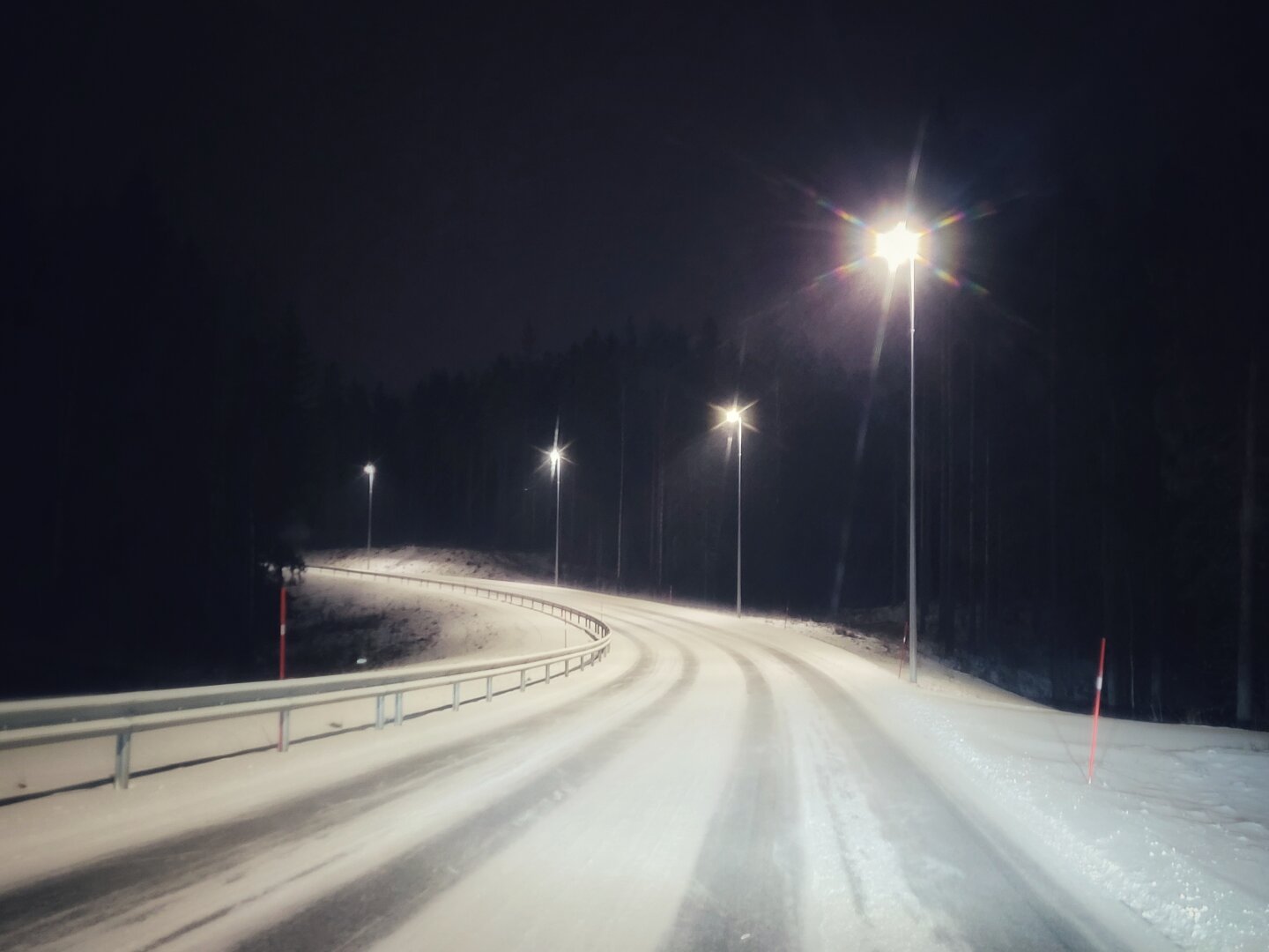 A road covered in snow, with lanes, winding its way through a dark forest, is lit up by streetlights.