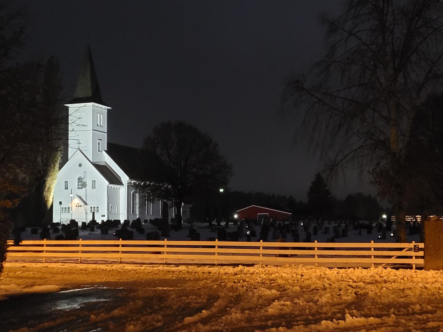 A white church, illuminated in the dark with spotlights, stands in the dark cemetery and appears to glow very white. The contrast is heightened by the fact that the wooden fence and the slush in front of it appear orange-yellow due to the streetlights