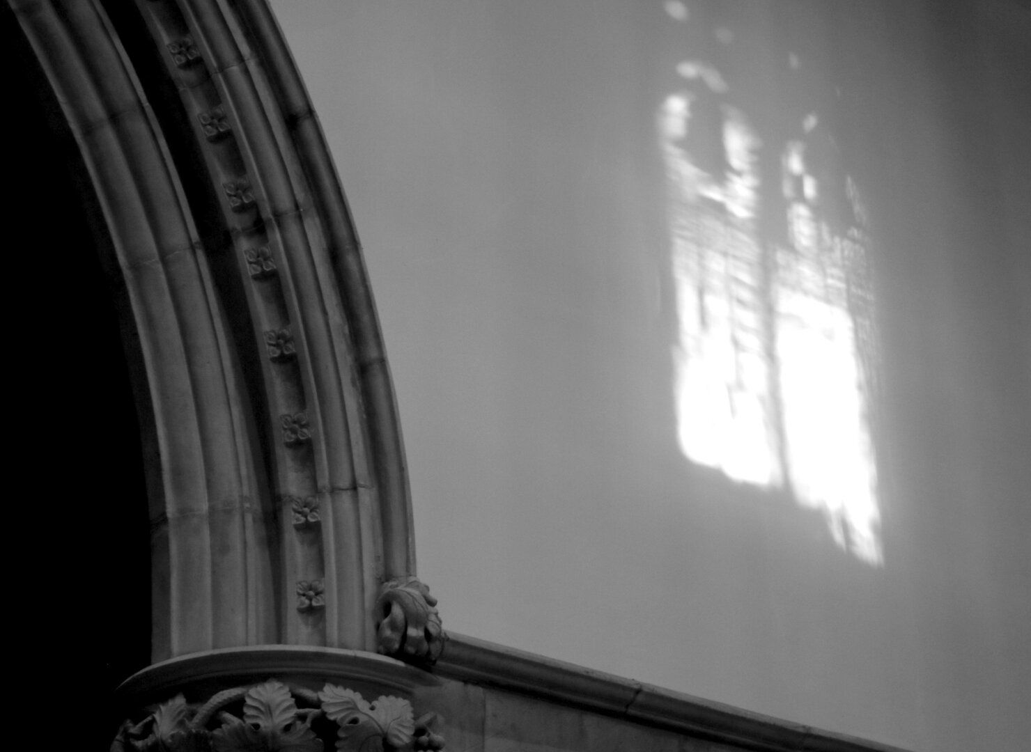 Black and white photography of sunlight projected on a wall, close and to the right of an arched frame decorated with carved flowers. The sun is coming through an unseen window on the opposite wall.