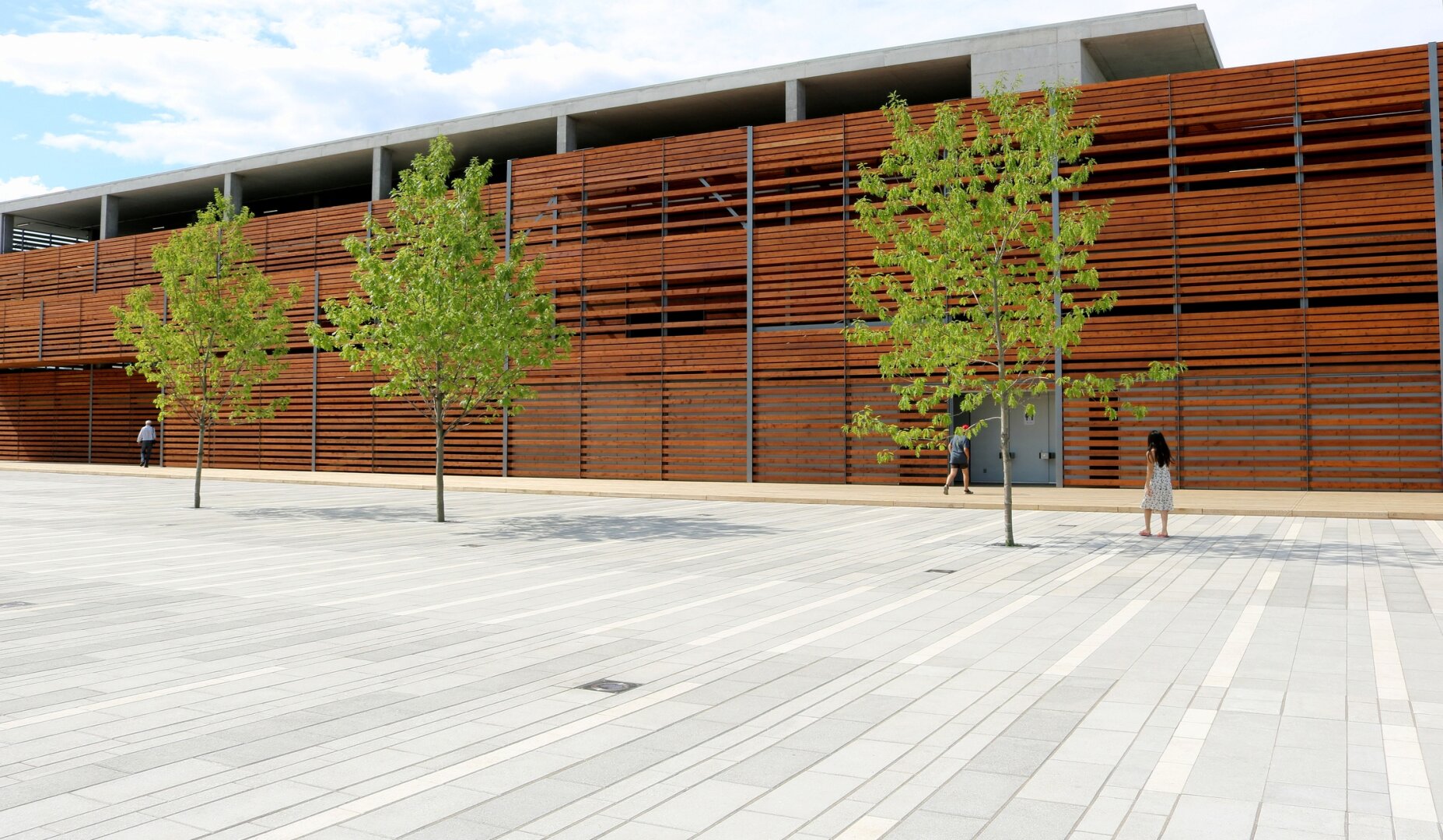 Photograph of a pale concrete building clad in horizontal, coffee-brown wooden slats. Gray rectangular paving stones form lines on the ground. Three tender green trees have been planted beneath the paving, as if they had no roots. The sunlight is very bright. A young girl with long black hair and wearing a sundress stands in the shade of one of the trees. Two other people walk on the sidewalk alongside the building. The scene is surreal and perfect, like an architect's model.

Photographie d'un bâtiment en béton pâle, recouvert de lattes horizontales de bois brun café. Au sol, des pavés rectangulaires gris forment des lignes. Trois arbres vert tendre ont été plantés sous le pavé, comme s'ils n'avaient pas de racines. La lumière du soleil est très éclatante. Une jeune fille aux longs cheveux noirs et à la robe soleil se tient dans l'ombre d'un des arbres. Deux autres personnes marchent sur le trottoir le long du bâtiment. L'ensemble est irréel et parfait, comme une maquette d'architecte