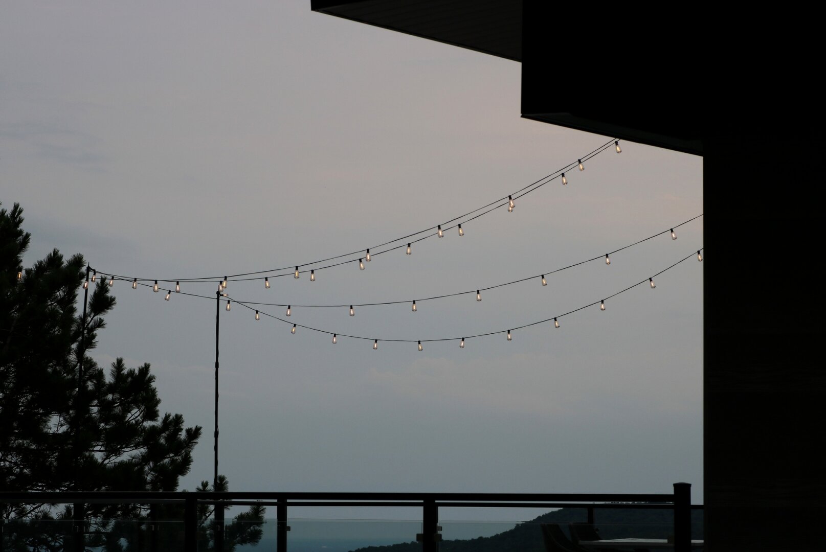 Photograph of a landscape surrounding the reception pavilion of an inn at the end of the day, showing a large enclosed patio above which four strings of lights are suspended, their bulbs lit and stretching across the image from left to right. To the left, the silhouette of tall pine trees. To the right, the silhouette of the building, with its straight lines. In the background, a sky with soft pinkish-blue hues, with a hill visible in the distance.

Photographie d'un paysage entourant le pavillon de réception d'une auberge à la fin du jour, montrant un grand patio clôturé au-dessus duquel sont suspendues quatre guirlandes de lumières, les ampoules allumées et traversant l'image de gauche à droite. À gauche la silhouette de grands pins. À droite, la silhouette du bâtiment, avec ses lignes droites. En arrière-plan, un ciel aux couleurs douces de bleu rosé, avec une colline visible au loin.