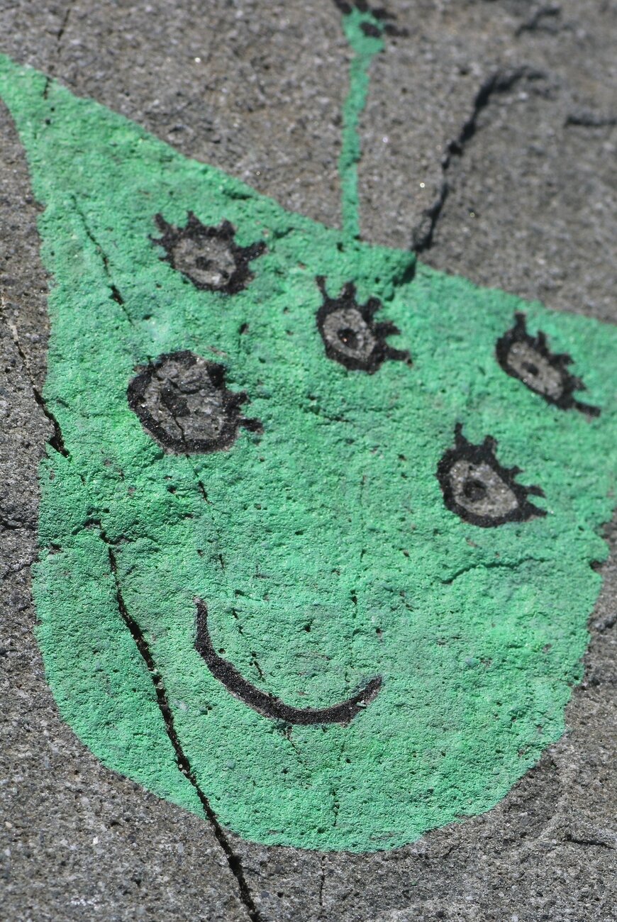 Photograph of a small turquoise monster painted on the gray surface of a rock, a simple rounded shape at the bottom and with two corners at the top, with a smile and five eyes with eyelashes, painted with black. The smiling creature also has a very straight antenna on top. The surface of the rock is rough and cracked.

Photographie d'un petit monstre turquoise peint sur la surface grise d'un rocher, une simple forme arrondie dans le bas et avec deux coins en haut, avec un sourire et cinq yeux avec des cils, peints avec du noir. La créature souriante a aussi une antenne bien droite sur le dessus. La surface de la roche est rugueuse et craquée.