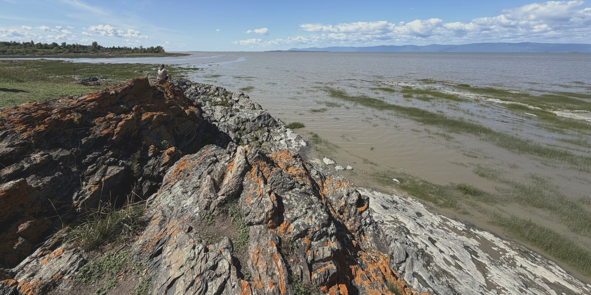 Photograph of a seaside landscape, showing in the foreground a grey and orange rocky cape advancing towards the sea and on which a woman is sitting, seen from behind. Green aquatic and shoreline plants then colonize the lower part of the shore, where the shallow water is slightly brownish. In the distance, the blue "sea" is actually the St. Lawrence River, already wide at this height, with the blue mountains of Charlevoix on the other side. On the left side, the mouth of the Port Joli River creates an opening in the bank.

Photographie d'un paysage de bord de mer, montrant à l'avant-plan un cap de roc gris et orange s'avançant vers la mer et sur lequel une femme est assise, de dos. Des plantes aquatiques et de rivage vertes colonisent ensuite la partie basse du rivage, où l'eau peu profonde est légèrement brunâtre. Au loin, la "mer" bleue est en réalité le fleuve St-Laurent, déjà large à cette hauteur, avec les montagnes bleues de Charlevoix de l'autre côté. Du côté gauche, l'embouchur