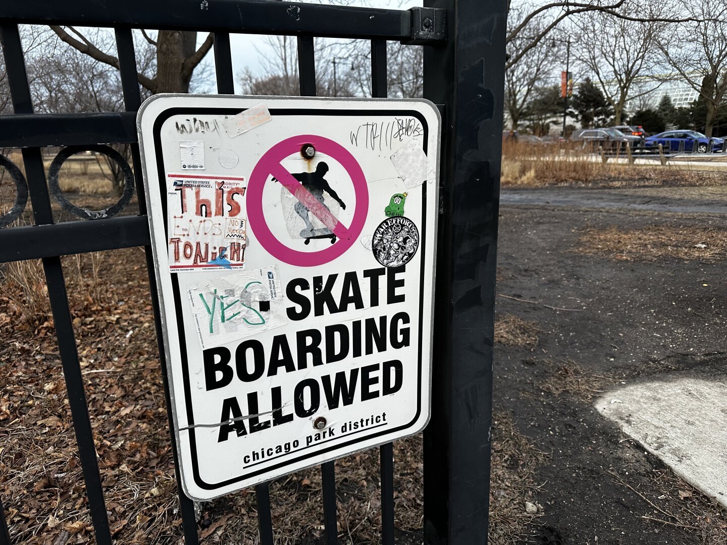 A city park sign on a fence is covered in stickers and graffiti. It reads “no skateboarding allowed,” but a “yes” sticker covers the “no.”