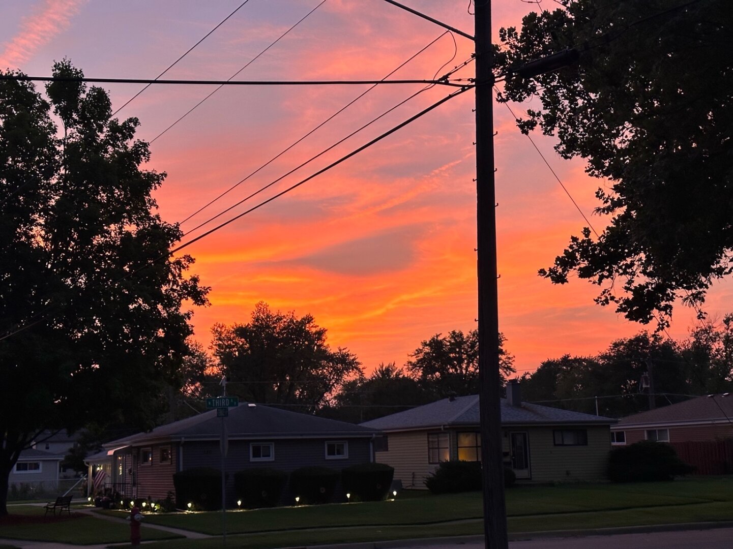 A house, trees and power poles are seen in silhouette against a dusk sky full of oranges and yellows and purples.