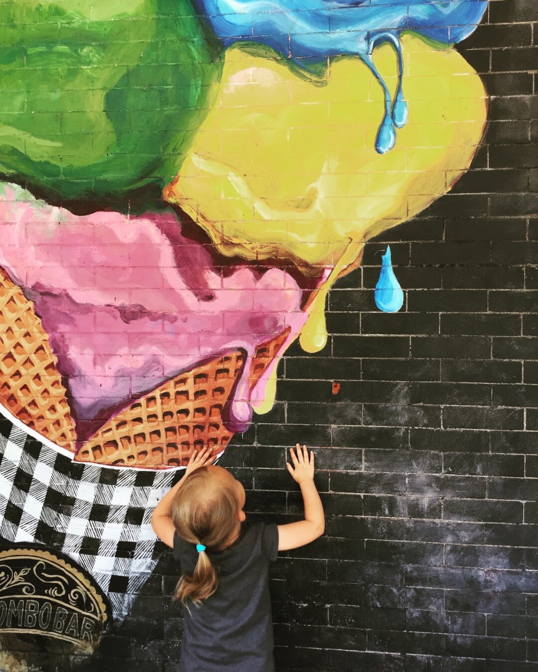 A young child with her back, turned to the camera, pretends to lick the ice cream depicted in a mural on a brick wall. ￼ The frozen desert is at least 12 feet tall, very drippy, and full of bright colors, such as yellow and pink. The ice cream is in a waffle cone wrapped in a black and white gingham napkin.