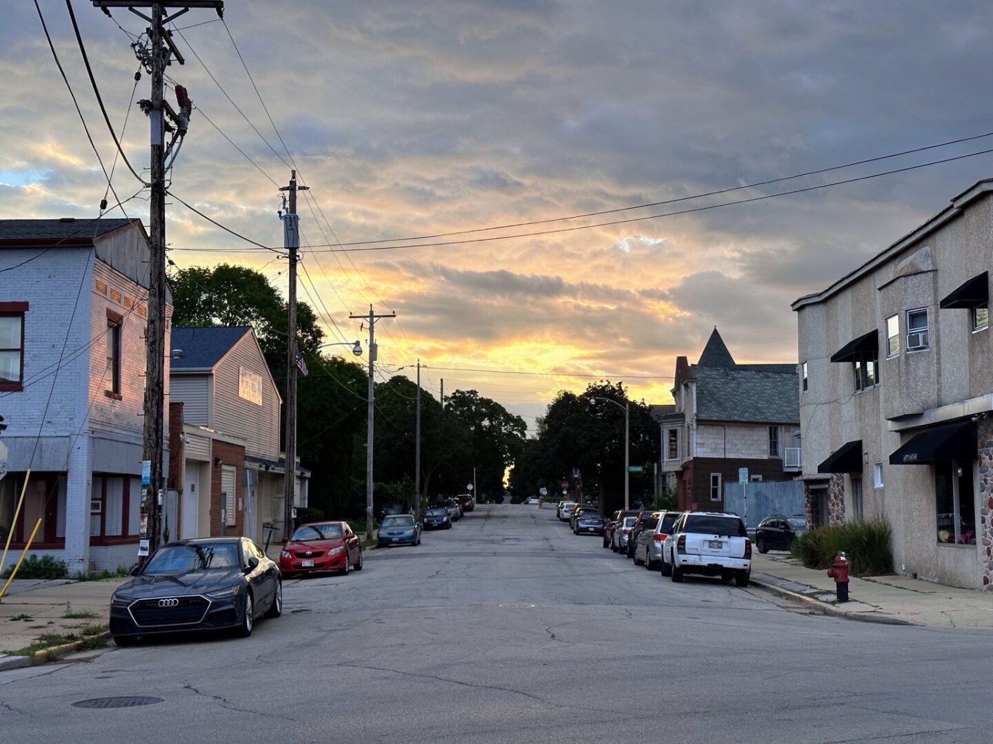 The sun sets far behind the trees down a blue collar residential street. The sky is cloudy and gray, but the sunset’s yellows and oranges highlight it.