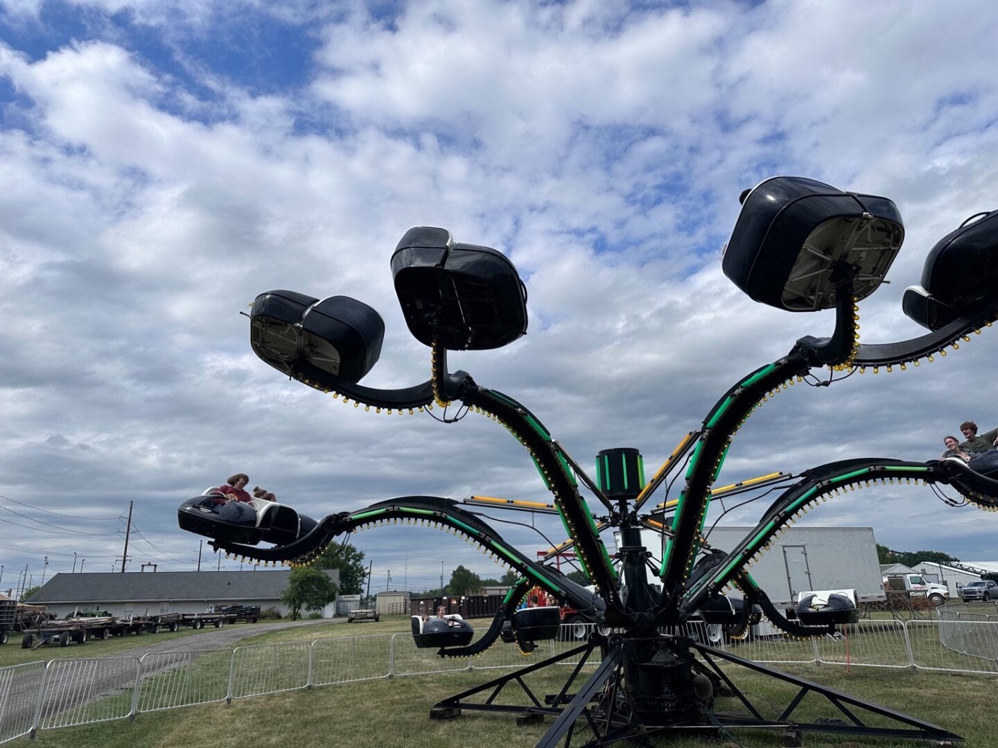 An octopus-themed carnival is in full operation, spinning a series of 16 black seats, some of which are tilted high in the air. The black machine presents in contrast to the blue sky full of white billowing cumulus clouds.