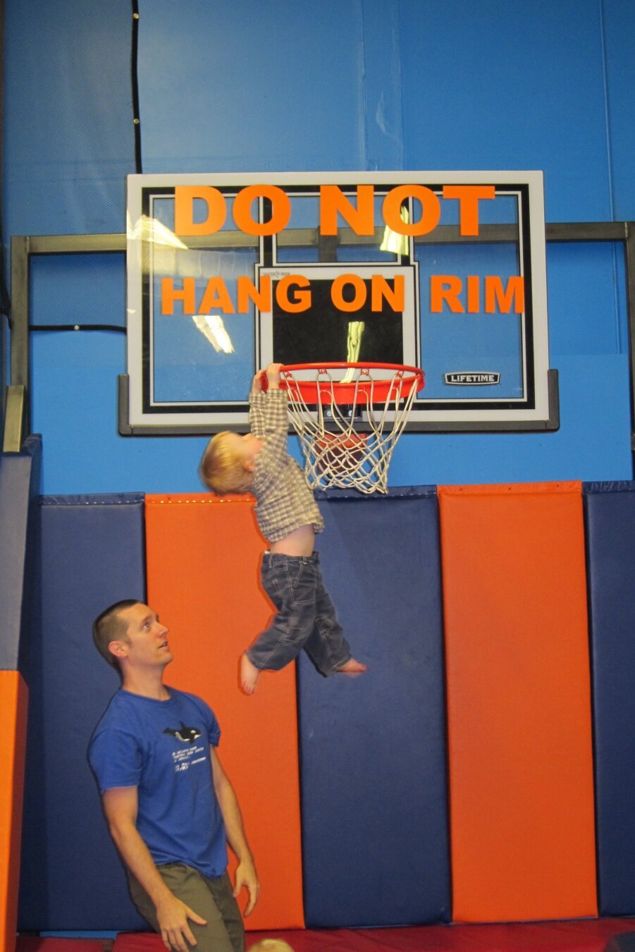 A toddler hangs from a basketball rim while his father looks on from nearby.  Large block text on the backboard reads "DO NOT HANG ON RIM".