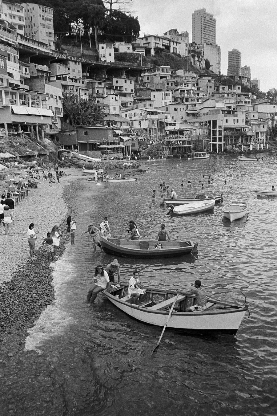 picture of gamboa favela in salvador, houses along cliffs with rowboats in water in foreground, many people on rocky beach