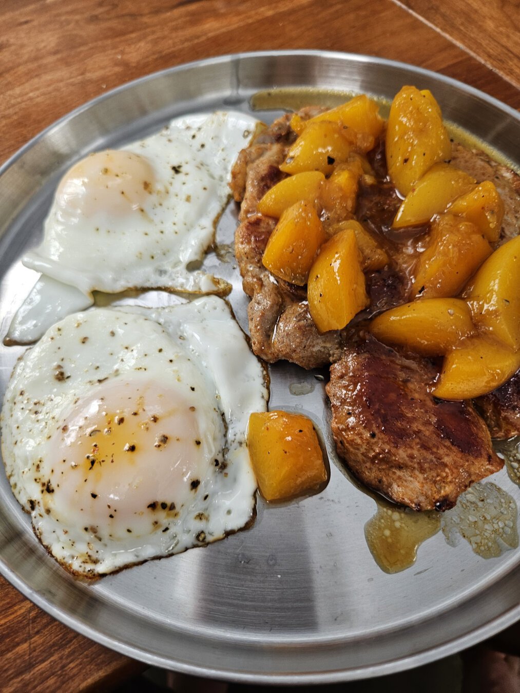 A metal plate of pork steaks topped with peach chutney and two eggs with flecks of fresh ground black pepper