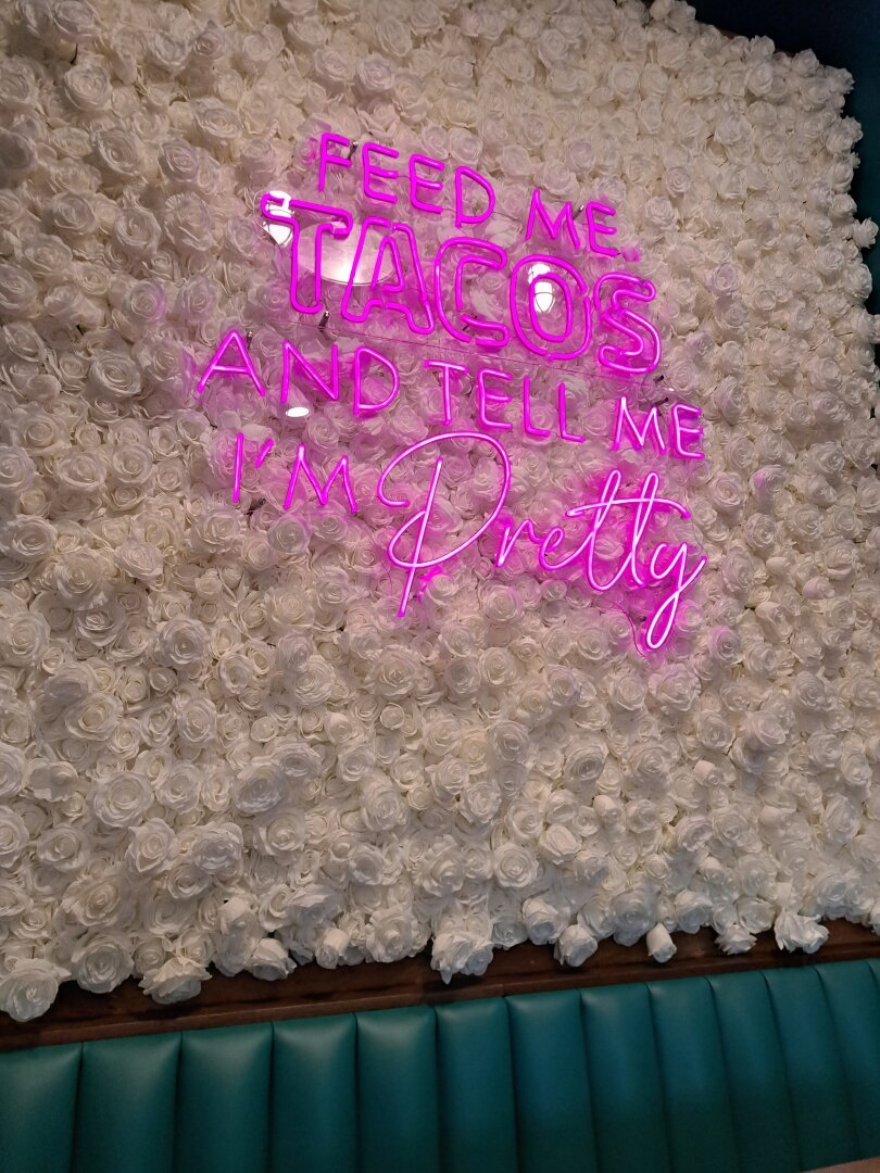 A sign in the restaurant is pink neon on white silk roses, saying: "Feed me tacos and tell me I'm pretty."